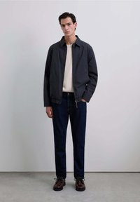 Chaqueta oscura con cuello, sobre un suéter claro. Jeans oscuros y zapatos marrones de cordones completan el conjunto. Tejido suave, corte relajado.