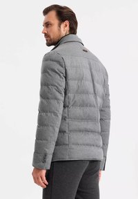 Chaqueta de plumas gris con diseño acolchado, cuello alto y puños abotonados. Textura suave con un brillo sutil. Detalles de costura notables en la parte posterior.