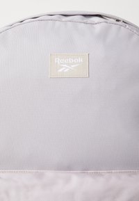 Mochila Reebok em cinza claro, feita de tecido texturizado. Apresenta uma etiqueta bege com logotipo branco, parte superior arredondada e fecho com zíper.