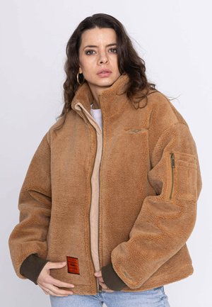 Femme aux longs cheveux foncés portant une veste polaire beige avec une fermeture éclair et des poignets marron, tenant le devant de la veste près de la taille.