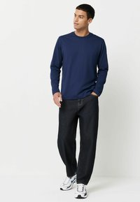 Cămașă lungă bleumarin cu manșete, asortată cu blugi largi de culoare închisă și adidași albi. Cămașa are un design clasic cu guler rotund și o textură netedă.
