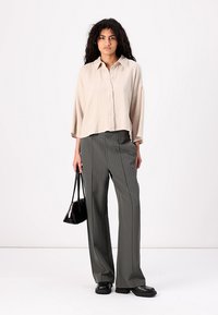 Bež srajca z gumbi in teksturirano tkanino, kombinirana s širokimi hlačami temno sive barve z navpičnimi črtami. Črna torbica in masivne črne čevlje.