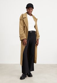 Cappotto trench beige doppiopetto con ampi revers, indossato sopra un maglione bianco. Abbinato a pantaloni neri a gamba larga e stivaletti neri chunky.