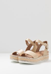 Beige Plattform-Sandalen mit Knöchelriemen, metallicenen Nieten auf gewebten Jute-Mittelsohlen und weißen Gummi-Außensohlen auf einer weißen Oberfläche.