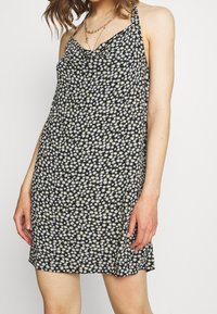 Robe noire sans manches avec un motif de marguerites blanches, dotée de fines bretelles et d'une coupe décontractée. Tissu léger avec une texture lisse.