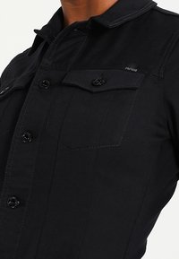 Czarna koszula denimowa z dwoma kieszeniami na piersi, zapięciami na guziki i kołnierzykiem. Posiada tonowane przeszycia i małe logo na piersi.