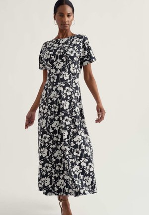 Kobieta ubrana w czarno-białą, kwiatową, maxi sukienkę z krótkim rękawem, stojąca z lekko odsuniętymi ramionami od ciała na jednolitym tle.