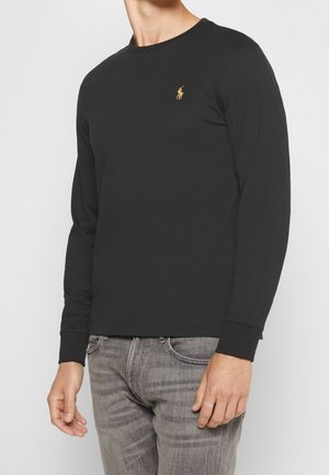 Man draagt een zwart shirt met lange mouwen en een klein goudkleurig geborduurd logo op de borst, gecombineerd met vervaagde grijze spijkerbroeken.