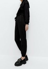 Persoon in zwarte joggingsbroek en een zwarte jas met elastische manchetten, gecombineerd met stevige zwarte loafers met decoratieve gespen.