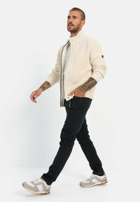 Knitted cream cardigan with a high collar and front buttons, paired with dark pants and grey sneakers, featuring textured fabric and two pockets.