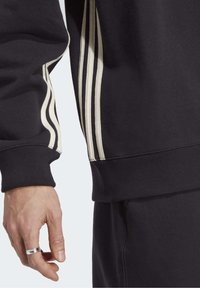 Svart sweatshirt med tre krämfärgade ränder längs ärmen och en ribbad manschett. Synlig hand med en silverring. Mjuk textur.