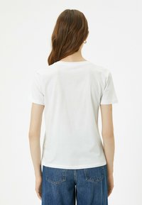 Ein weißes Baumwoll-T-Shirt mit kurzen Ärmeln und rundem Halsausschnitt, das eine glatte Textur aufweist. Trägt dazu eine Jeans aus Denim und ist von hinten zu sehen.