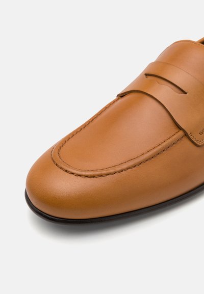 Tan leather loafer with a smooth finish, rounded toe, stitched detailing along the edge, and a decorative strap across the vamp.