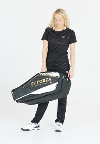 Svart sportoutfit med en texturerad design, håller en stor FZ Forza sportväska med svart och vit färg och guldlogodetaljer.