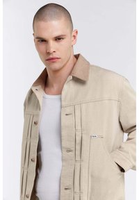 Giacca in denim beige con colletto marrone, dotata di bottoni di chiusura, due tasche laterali e una piccola etichetta del marchio sulla tasca sinistra.