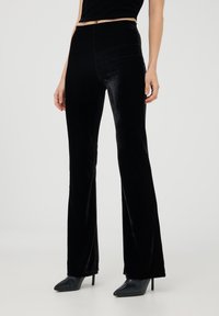 Pantaloni a zampa d'elefante in velluto nero con una texture liscia e alta vita. Presentano una silhouette aderente che allunga la linea delle gambe, abbinati a tacchi a punta.