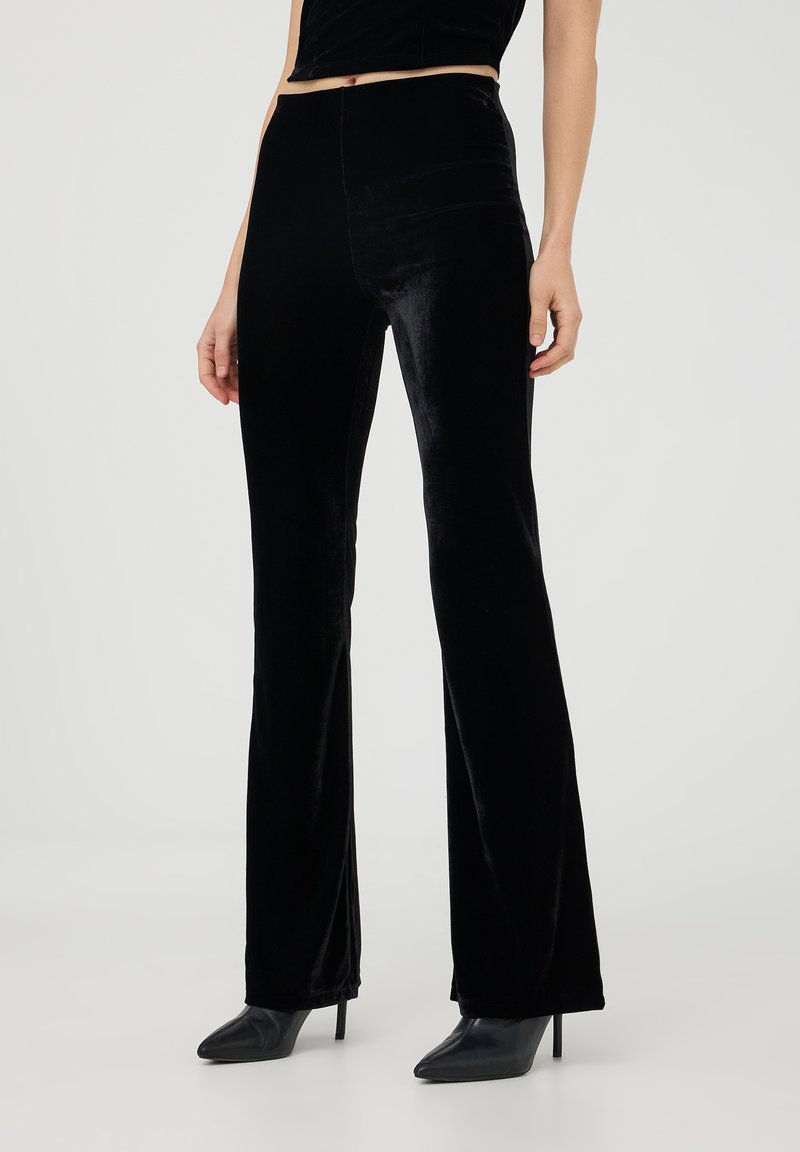 Pantaloni a zampa d'elefante in velluto nero con una texture liscia e alta vita. Presentano una silhouette aderente che allunga la linea delle gambe, abbinati a tacchi a punta.