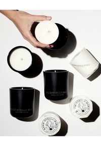 Schwarze Kerzenbehälter mit weißem Wachs, die mit geprägtem Text ausgestattet sind. Eine Hand hält eine weiße Kerze. Daneben befinden sich zwei runde weiße Pakete.