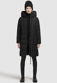 Černá puffer bunda s kapucí, s obousměrným zipem, prošívaným designem a bočními kapsami. Nositá přes černý rolák a legíny.