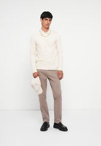 Langarmige crèmekleurige polo met kraag, beige broek, zwarte loafers en een lichtgrijze pet in de hand. Eenvoudig ontwerp en neutrale kleuren.