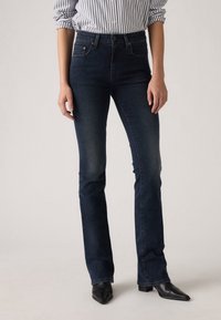 Femme portant un jean bootcut bleu foncé, une chemise à manches longues rayée avec les poignets retroussés, et des bottines noires à bout pointu, debout sur un fond neutre.
