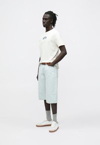 Hellfarbenes T-Shirt mit grafischem Druck, hellblaue Jeansshorts, graue Socken und weiße Sneakers mit braunen Akzenten, stehende Pose.