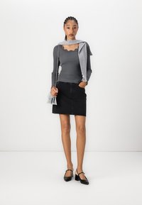 Haut gris à manches longues avec un col festonné, associé à une jupe en denim noire. Le mannequin tient un petit sac à main gris et porte des talons noirs.