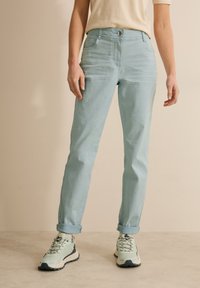 Jeans de color azul claro con un ajuste ligeramente entallado, que cuentan con bolsillos frontales, cierre de botón y puños doblados. Combinados con zapatillas de color verde claro.