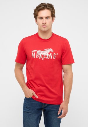 Junger Mann trägt ein rotes Mustang-T-Shirt mit einem weißen Pferde-Motiv und dunkelblaue Jeans, steht mit einer Hand in der Tasche.