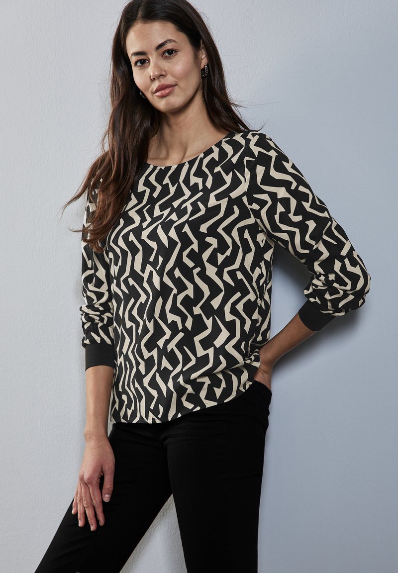 Blusa de manga larga en negro y crema con un patrón de zigzag. Presenta un escote redondo y puños elásticos. Está confeccionada con una tela suave y ligera.