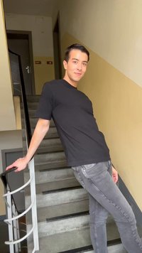 Νέος άνδρας με μαύρο μπλουζάκι και γκρι τζιν, ακουμπισμένος στη κουπαστή της σκάλας μέσα σε ένα κτίριο με μπεζ τοίχους, χαμογελώντας στην κάμερα.