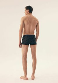 Czarne kąpielówki w sześciokątny wzór, dopasowany krój i gładka faktura materiału, noszone przez osobę widzianą od tyłu.