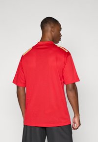 Piros hálós sportmez rövid ujjakkal és gallérral. Arany díszítéssel a vállakon. Texturált anyag a légáteresztés érdekében.