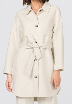 Femme portant un manteau crème ceinturé avec des boutons à l'avant et des poches poitrine, debout devant un fond clair uni.