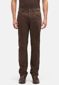 Mężczyzna w brązowych spodniach o prostych nogawkach i dopasowanych brązowych skórzanych butach, stojący na tle jednolitego, jasnego tła.