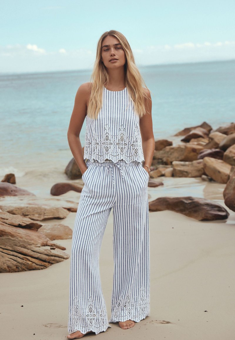 Femme debout pieds nus sur une plage de sable près des rochers, vêtue d'un haut sans manches blanc et bleu à rayures et d'un pantalon à jambes larges avec des détails en œillets.