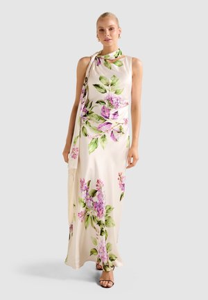 Ženska v kremni maxi obleki brez rokavov s purpurno-zelenim cvetličnim vzorcem, kombinirana z ujemajočo se cvetlično rutko in zlatimi obročastimi uhanami.