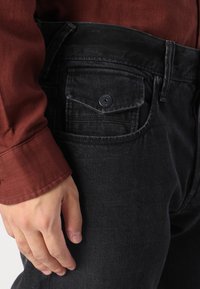 Jeans din denim negru cu o croială structurată, având o buzunar frontal cu închidere prin buton și o textură subtilă. Mână odihnindu-se pe șold.