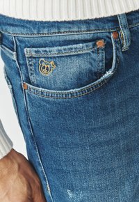 Vaqueros de mezclilla con un acabado azul deslavado, que presentan un logo de oso bordado en el bolsillo trasero y detalles en botones de tono cobre.