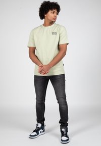 Svetle zelené bavlněné tričko s kulatým výstřihem a malým černým logem na hrudi, doplněné o tmavě šedé skinny džíny a tenisky s černými akcenty.