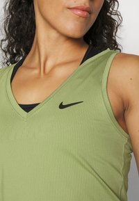 Kobieta nosząca zielony bezrękawnik sportowy Nike z czarnym stanikiem sportowym pod spodem, pokazująca ramię, szyję i część twarzy.