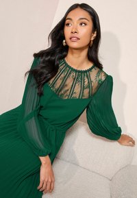 Rochie verde cu maneci lungi, cu un guler din dantela transparenta cu modele florale. Fusta plisata, materialul are o textura neteda. Modelul este asezat pe o canapea deschisa.