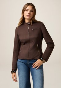 Veste marron zippée avec un col montant, poches latérales zippées et une poche poitrine. Texture lisse avec un matériel minimal et un design ajusté.