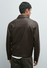 Man met kort, donker haar, gekleed in een donkerbruine leren jas en donkere jeans, met zijn rug naar de camera tegen een effen lichte wand achtergrond.