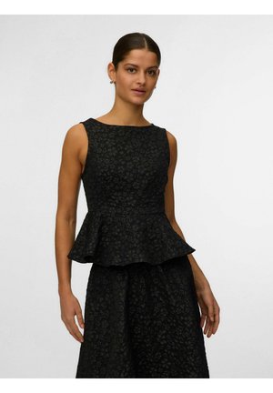 Femeie cu păr închis la culoare, purtând un top negru fără mâneci cu model floral și volănaș, și o fustă asortată, stând pe un fundal simplu, deschis la culoare.