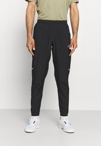 Schwarze Sporthose mit lockerer Passform, elastischem Bund, seitlichen Taschen und reflektierenden Akzenten. Kombiniert mit weißen Turnschuhen.