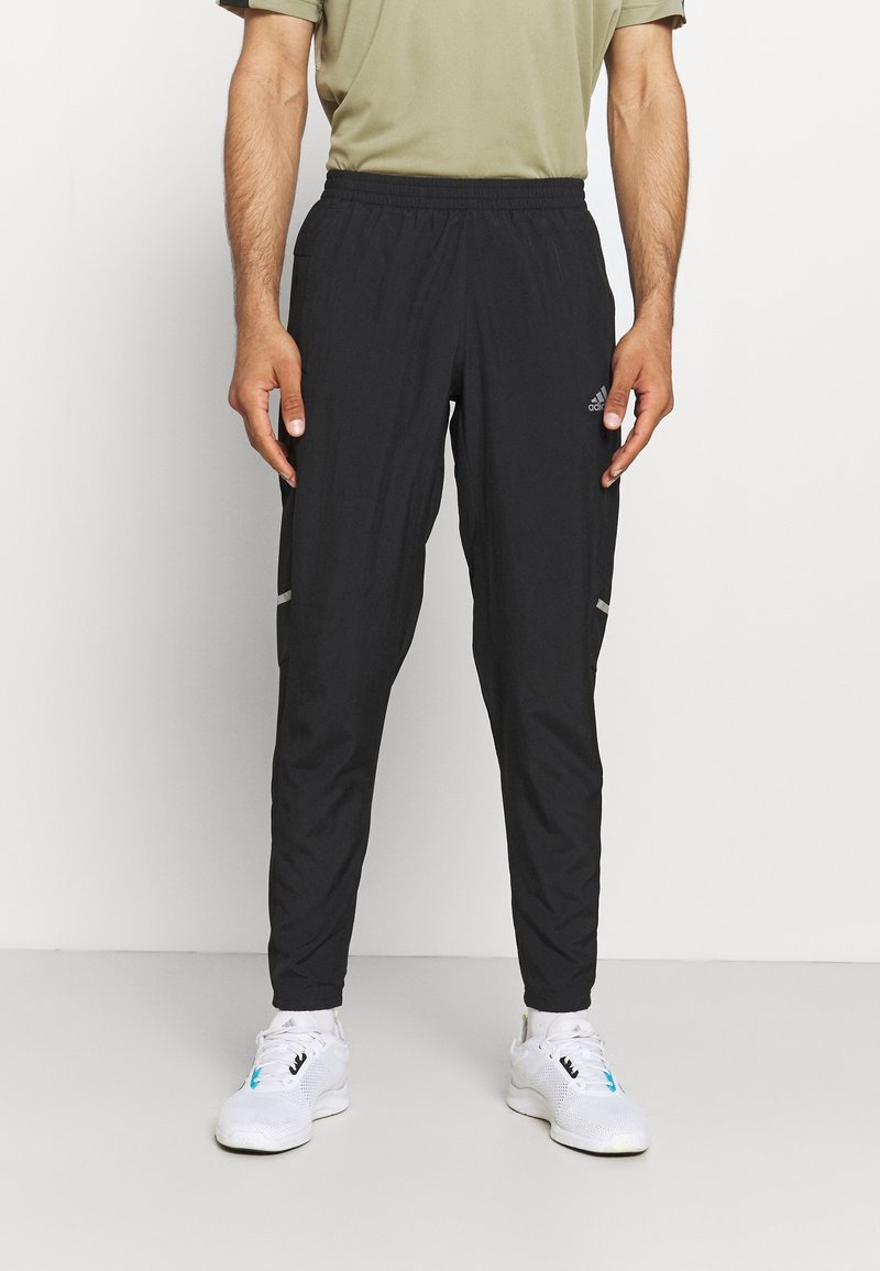 Schwarze Sporthose mit lockerer Passform, elastischem Bund, seitlichen Taschen und reflektierenden Akzenten. Kombiniert mit weißen Turnschuhen.