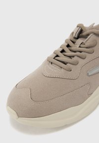 Beige suede sneaker with rounded toe, textured surface, subtle stitching details, and cream-colored rubber sole. Laces are flat and matching.