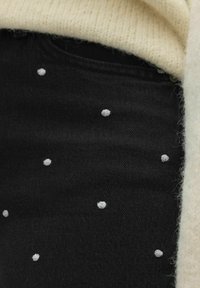 Svart denimstoff med små hvite prikkemønstre. Et lyst, ribbestrikket stoff titter frem fra den øverste kanten. Teksturert overflate synlig.