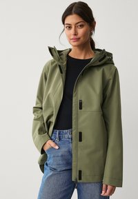 Chaqueta impermeable color caqui con capucha, con herrajes negros y puños ajustables. Combinada con una camiseta negra y jeans de mezclilla azules.